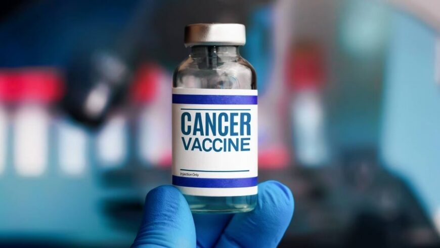 ​صورة مقربة لقارورة طبية (Vial) مكتوب عليها بوضوح "CANCER VACCINE" (لقاح السرطان) يحملها شخص يرتدي قفازات طبية زرقاء، ترمز للتطورات المخبرية في علاج الأورام.