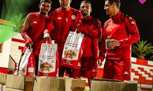 لاعبو نادي الوداد الرياضي يرتدون الملابس الرياضية الحمراء ويقومون بتعبئة كراتين المساعدات الغذائية "قفة رمضان" فوق أرضية الملعب ليلاً.