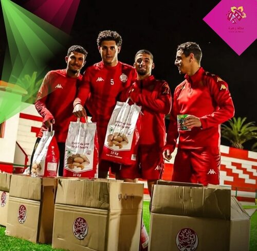 لاعبو نادي الوداد الرياضي يرتدون الملابس الرياضية الحمراء ويقومون بتعبئة كراتين المساعدات الغذائية "قفة رمضان" فوق أرضية الملعب ليلاً.