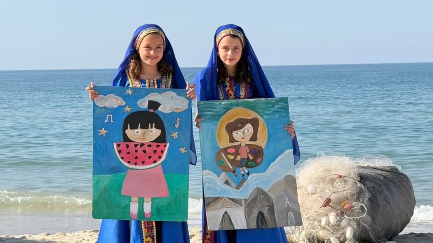فتاتان بالزي الشعبي الفلسطيني على الشاطئ تحملان لوحات رسم يدوية، إحداهما تضم رسمة لبطيخة والأخرى تجسد الخيام، ضمن مبادرة فنية في غزة.